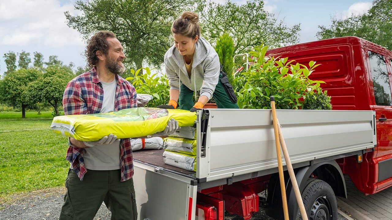 Dvoje ljudi istovaruje biljke i vreće sa zemljom iz crvenog Volkswagen Craftera. Scena se odvija na otvorenom, sa zelenilom i drvećem u pozadini. Atmosfera je radna i prirodna.