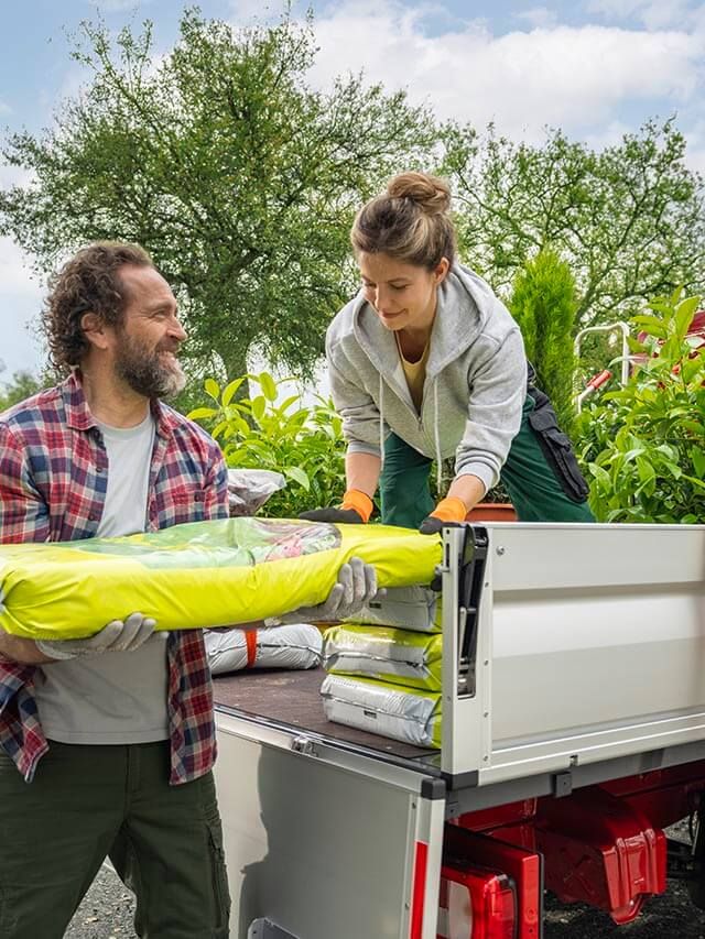 Dvoje ljudi istovaruje biljke i vreće sa zemljom iz crvenog Volkswagen Craftera. Scena se odvija na otvorenom, sa zelenilom i drvećem u pozadini. Atmosfera je radna i prirodna.