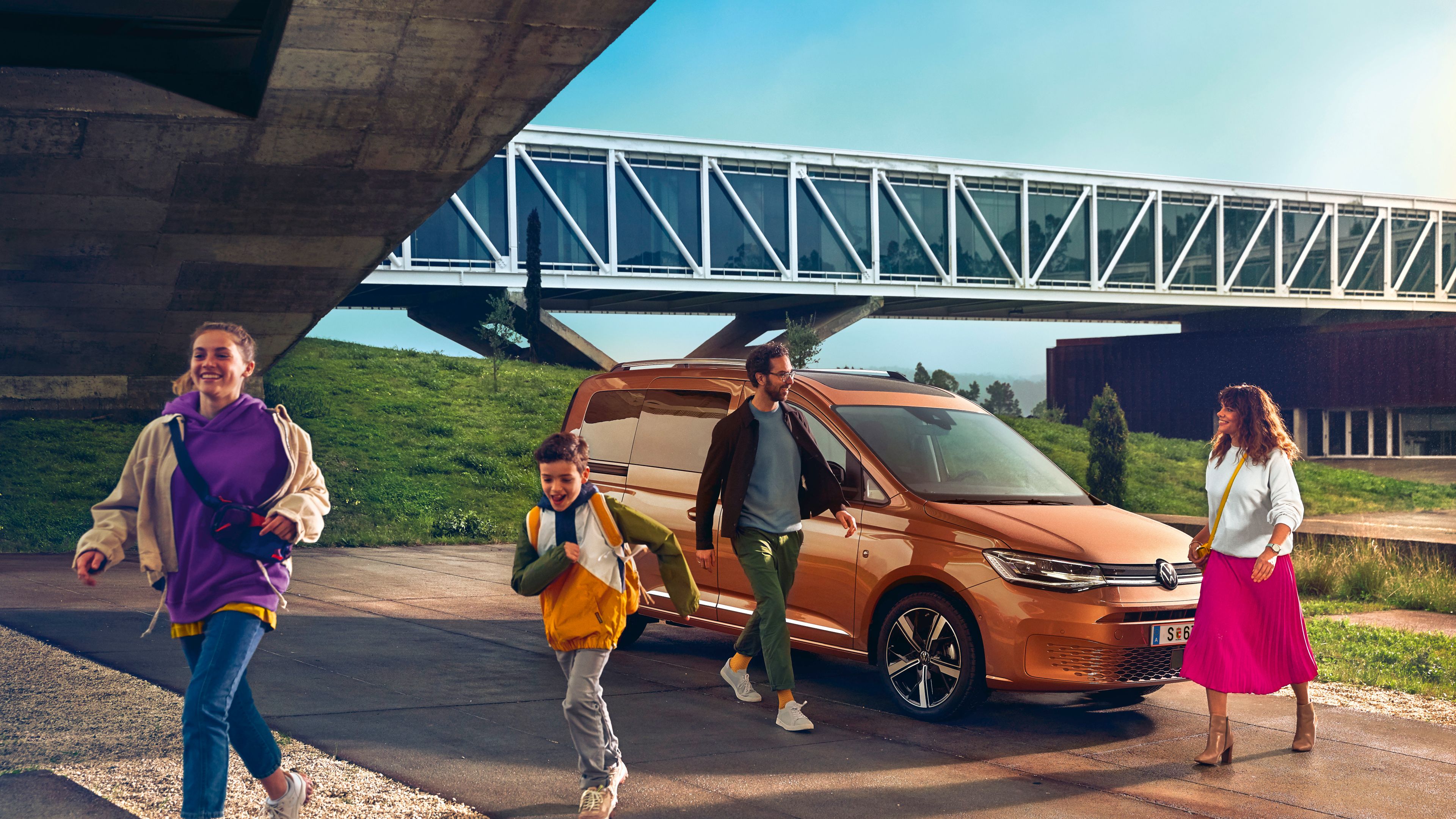 Ein VW Caddy 2020 parkt vor einer Brücke. Eine Familie geht gerade vom Auto weg.