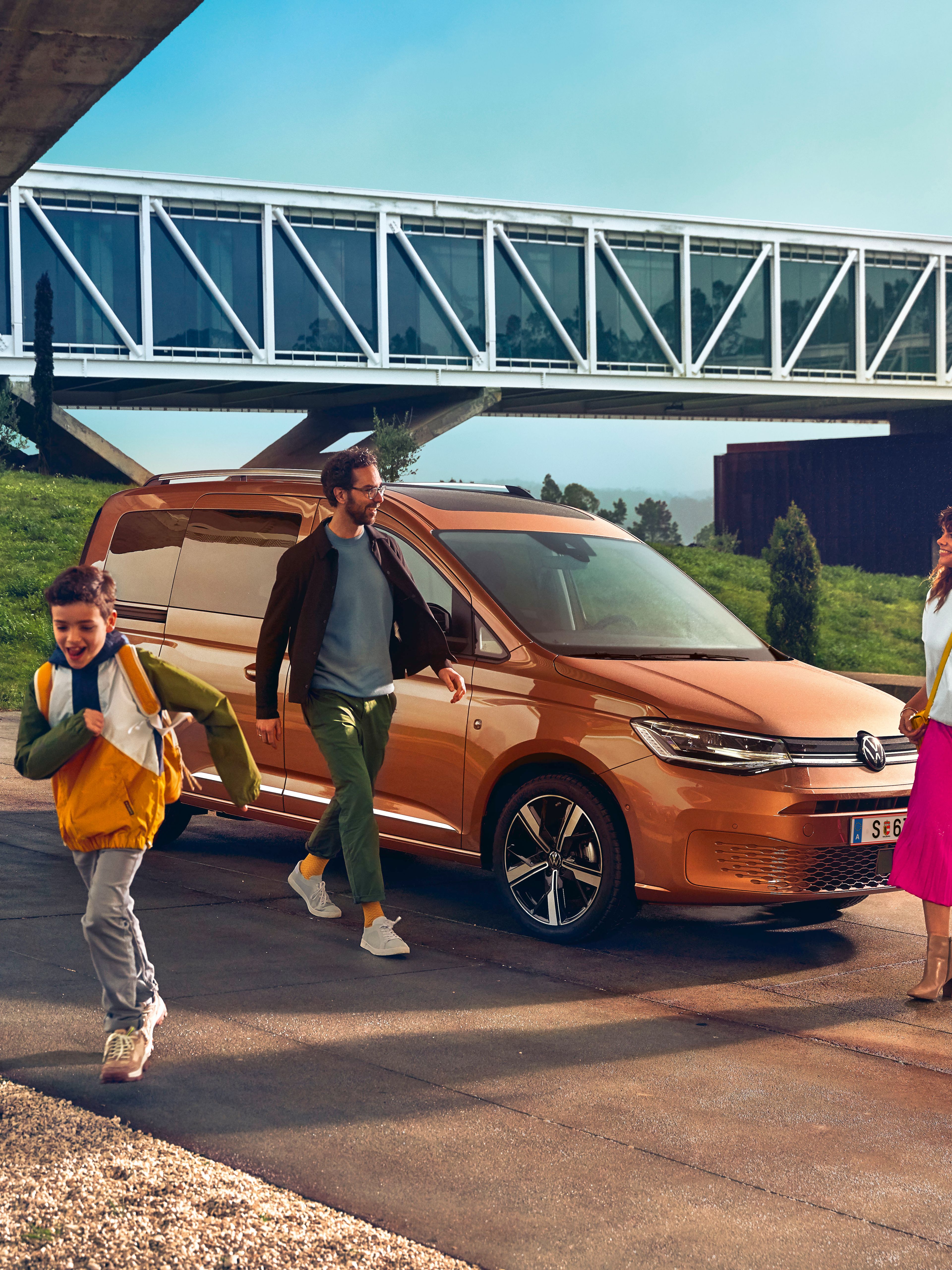 Ein VW Caddy 2020 parkt vor einer Brücke. Eine Familie geht gerade vom Auto weg.
