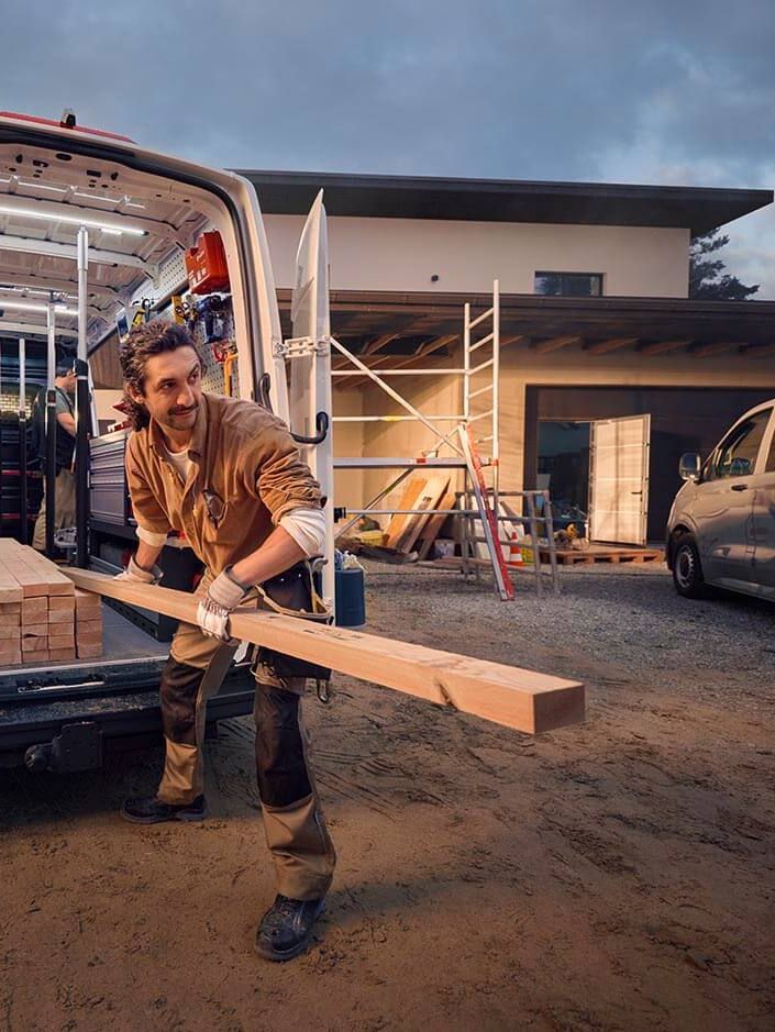 Radnik istovara drvene daske iz Volkswagen Crafter u blizini kuće u izgradnji. U pozadini su vidljivi alat i drugi materijali neophodni za građevinske radove.