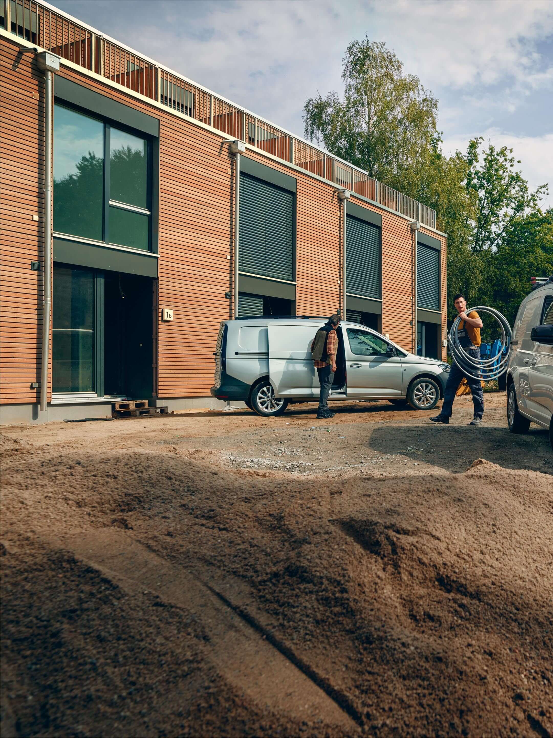 Ljudi rade na građevinskom mjestu ispred moderne zgrade sa bijelim Volkswagen Caddy punim opreme. Jedna osoba nosi kovčeg sa alatima, dok druga osoba pridržava električne kablove. Ispred kombija nalazi se gomila pijeska.