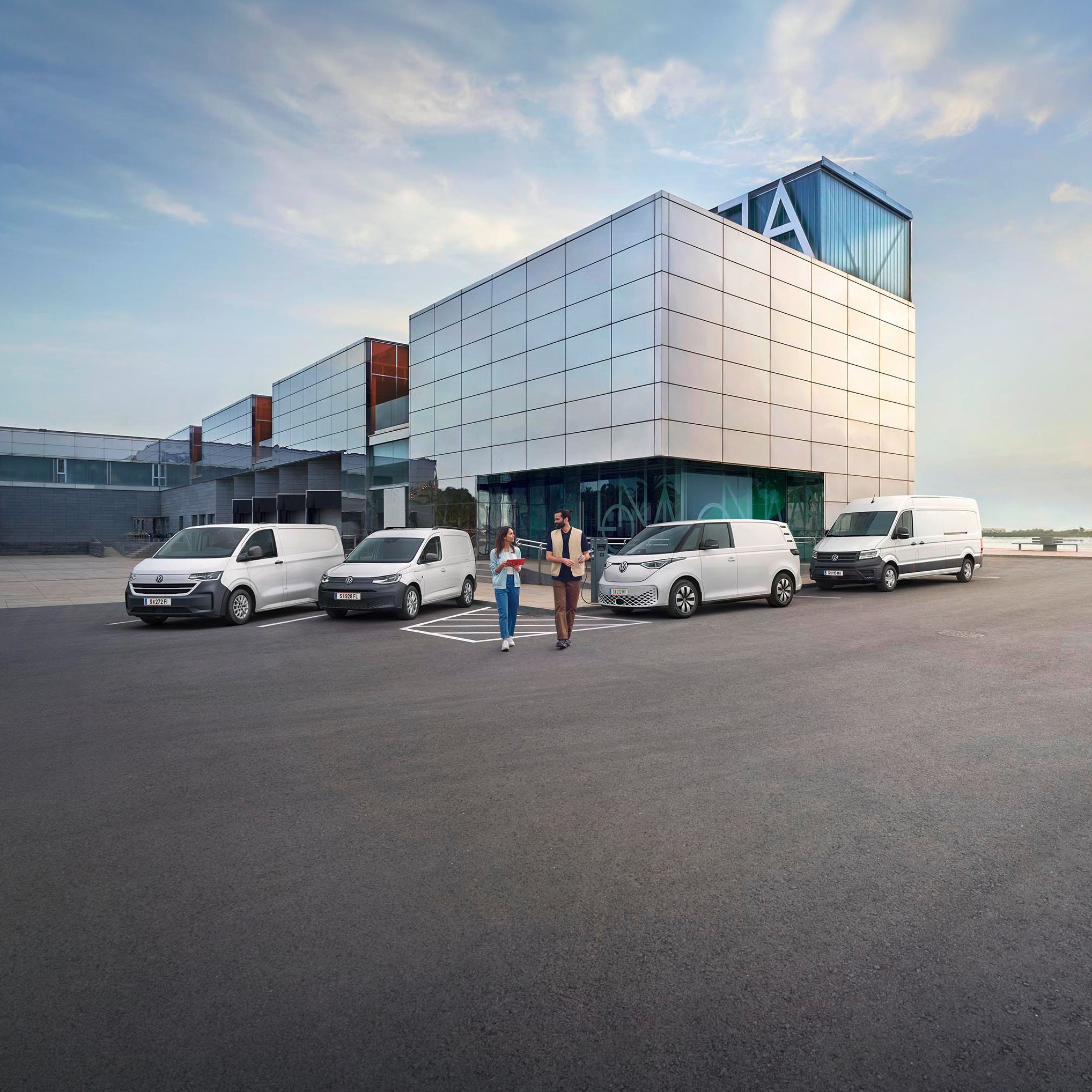 Ein modernes Lagergebäude mit Verladebereichen und mehreren geparkten Transportern von Volkswagen Nutzfahrzeuge. Zwei Personen stehen vor dem Gebäude und unterhalten sich. Der Himmel ist teilweise bewölkt, und es gibt einen Baum in der Ferne.