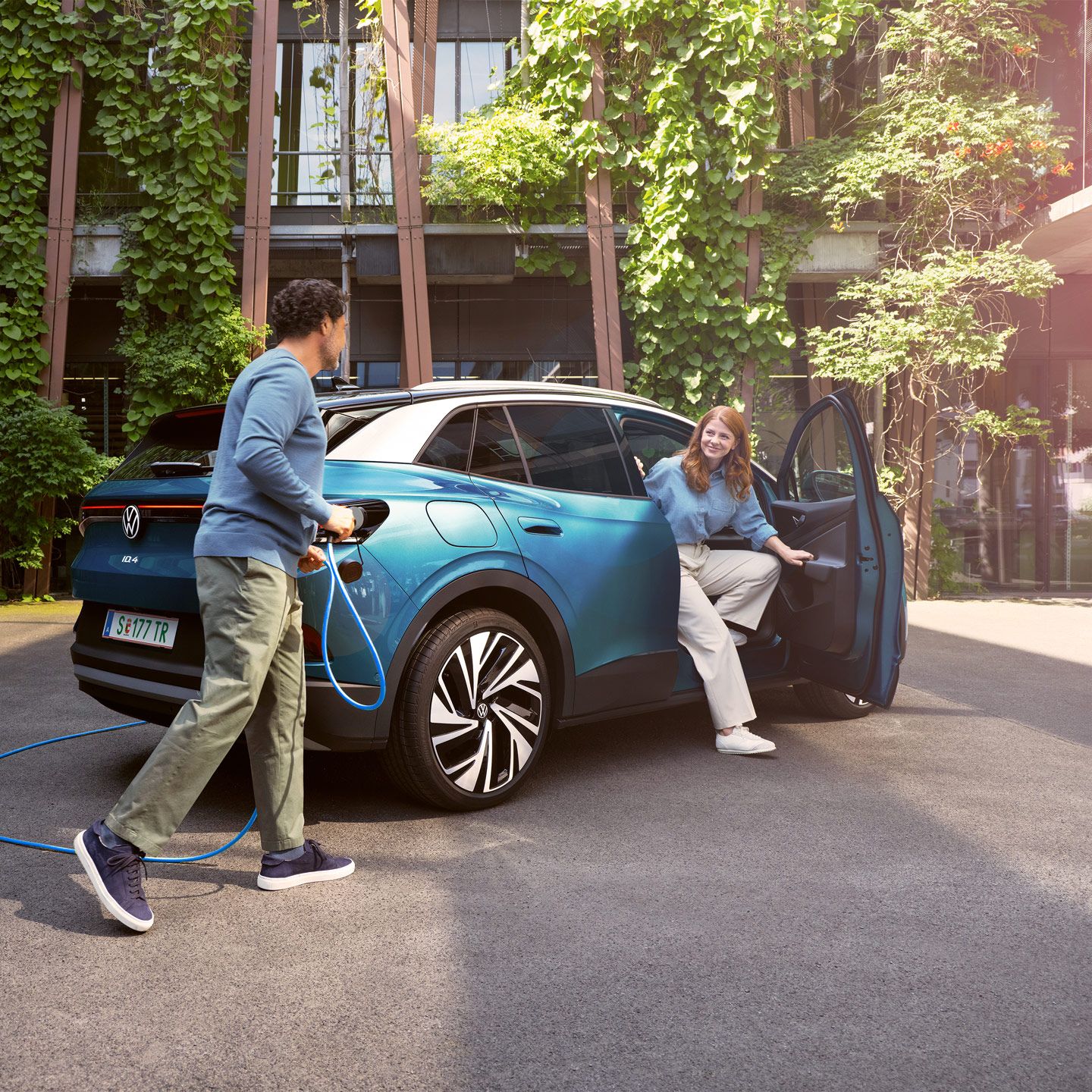 Eine Frau steigt aus einem blauem ID.4 aus, während ein Mann das POWER 2 Go Kabel anschließt. Im Hintergrund sind grüne Pflanzen und moderne Gebäude zu sehen.