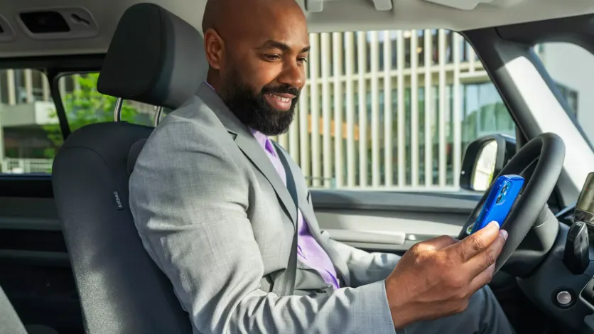 Ein Mann in einem grauen Anzug sitzt in einem Auto und schaut auf sein Smartphone. Sein Gesicht ist unscharf gemacht. Im Hintergrund sind moderne Gebäude zu sehen.