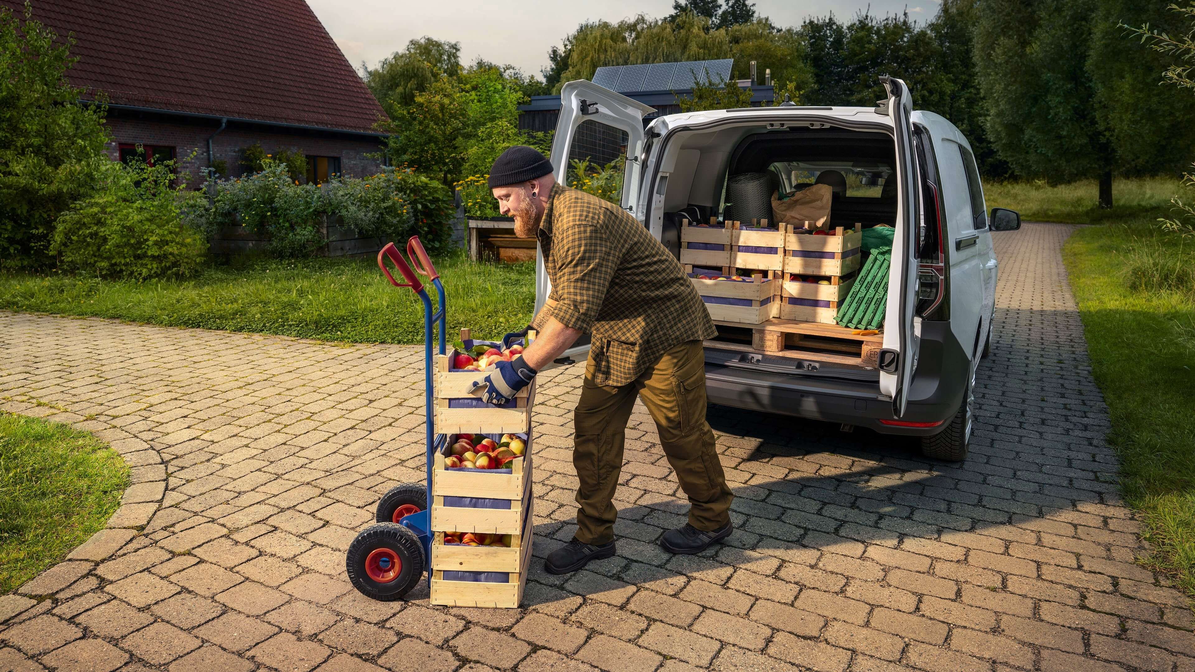 Osoba istovara drvene sanduke sa sezonskim voćem i povrćem iz bijelog Volkswagen Caddy parkiranog na seoskom imanju. Sanduci su postavljeni na ručna kolica. Okruženje je zeleno s mnoštvom vegetacije i kućama u pozadini.