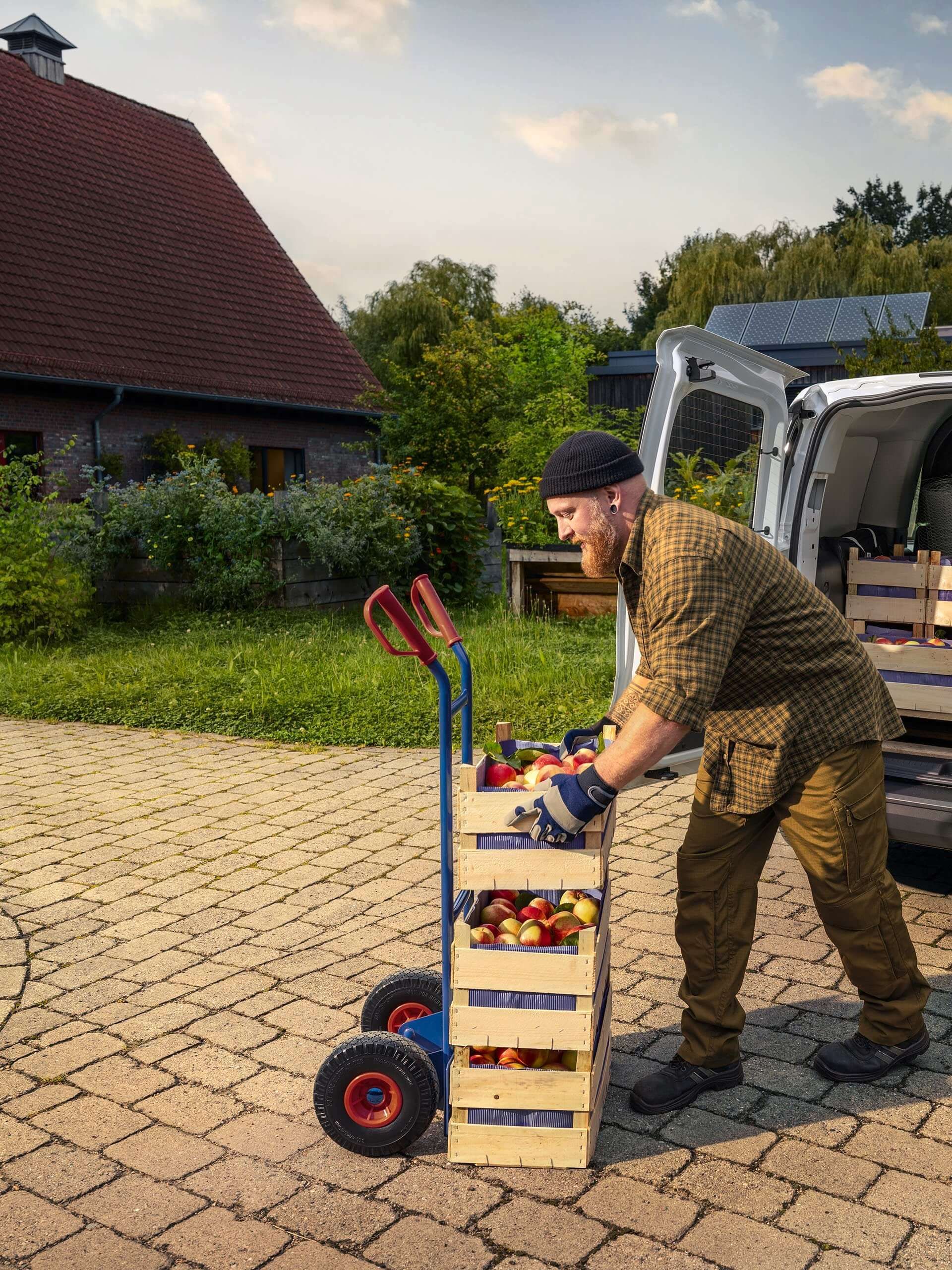 Osoba istovara drvene sanduke sa sezonskim voćem i povrćem iz bijelog Volkswagen Caddy parkiranog na seoskom imanju. Sanduci su postavljeni na ručna kolica. Okruženje je zeleno s mnoštvom vegetacije i kućama u pozadini.